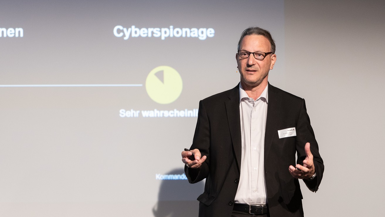 Ein Mann im dunklen Anzug spricht auf einer Bühne vor einer Leinwand mit der Aufschrift „Cyberspionage“ und einer Grafik. Er trägt eine Brille und ein Headset-Mikrofon, gestikuliert mit beiden Händen und wirkt engagiert, während er seine Präsentation erläutert.