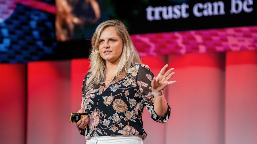 Eine blonde Frau steht auf einer rot beleuchteten Bühne und spricht zum Publikum. Sie trägt eine dunkle Bluse mit Blumenmuster, hält einen Presenter in der linken Hand und gestikuliert mit der rechten.