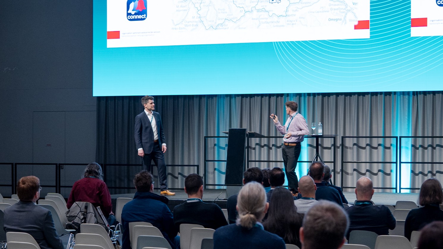 Auf einer Konferenzbühne halten zwei Männer vor einer grossen, hellblauen Leinwand eine Präsentation. Davor sitzt das Publikum auf Stühlen und schaut zu. Der Blickwinkel ist von den hinteren Reihen aus.