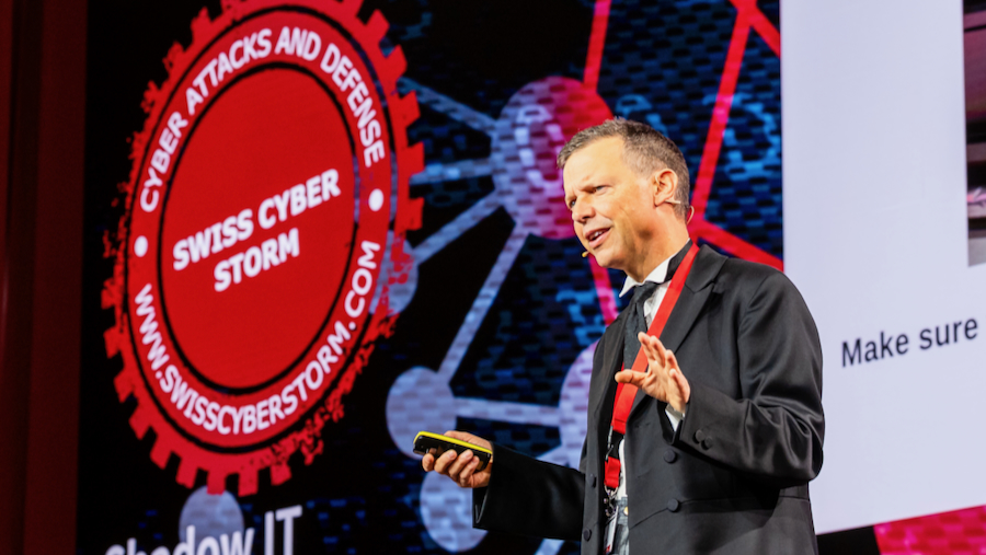 Ein Mann im dunklen Anzug spricht auf einer Bühne und gestikuliert mit der rechten Hand. Hinter ihm ist auf einer grossen Leinwand das rot-schwarze Logo der Swiss Cyber Storm zu sehen.