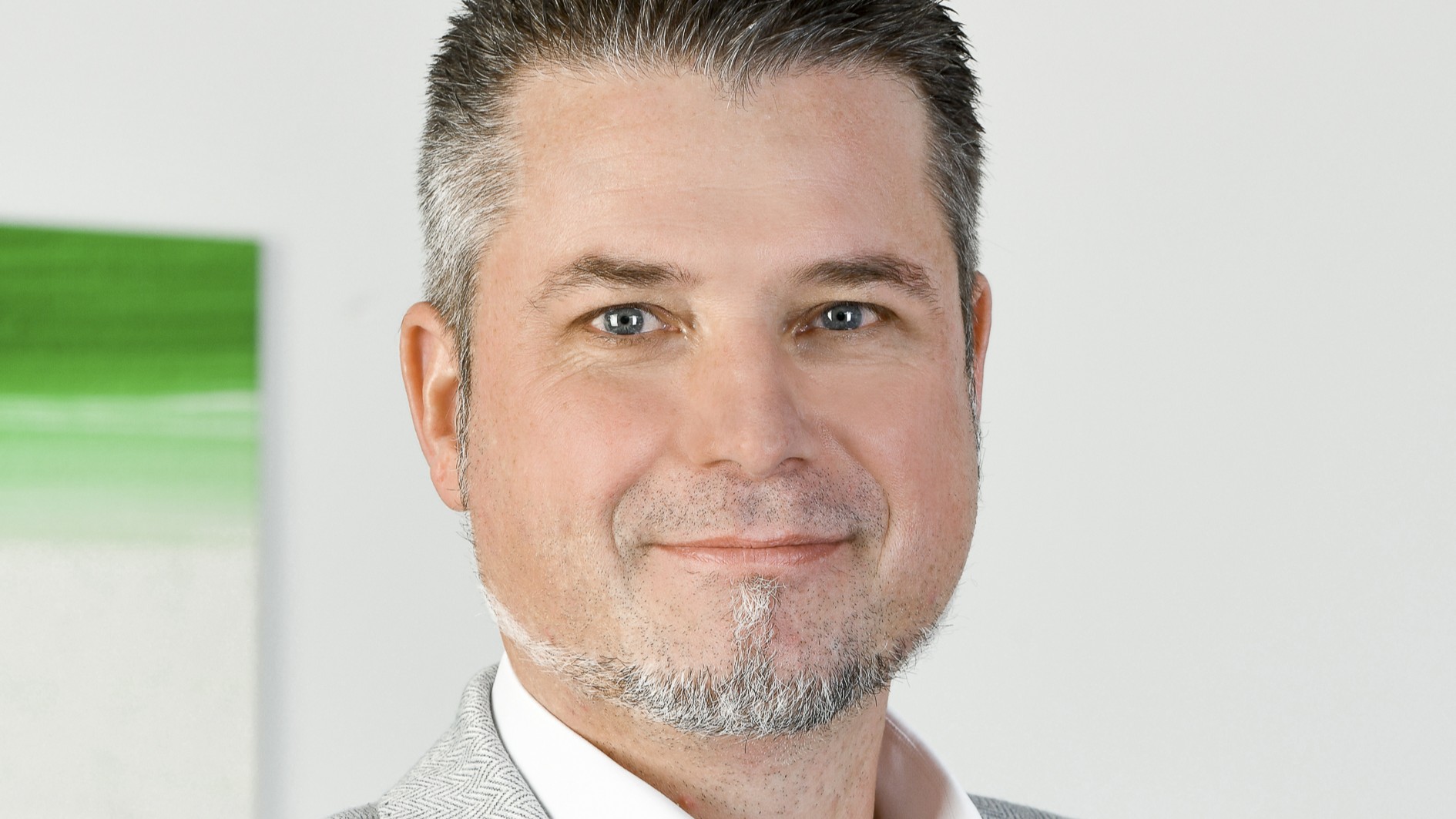 Porträt eines freundlich lächelnden Mannes mittleren Alters vor hellem Hintergrund. Er blickt direkt in die Kamera, hat blaue Augen, kurzes, grau meliertes Haar und einen gepflegten grauen Spitzbart. Er trägt ein helles Sakko über einem weissen Hemd.