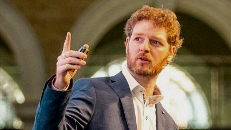 Robert McArdle hält eine Präsentation und gestikuliert im Bild. Dabei hält er eine Fernbedienung für eine Präsentation in der Hand.