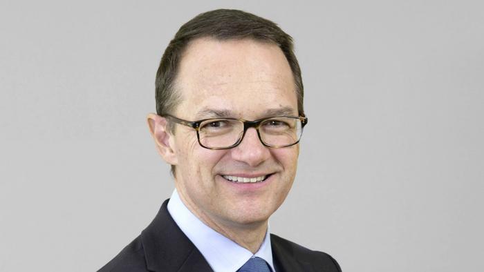 Mann mittleren Alters in dunkelblauem Anzug mit Lesebrille vor neutral-grauem Hintergrund.