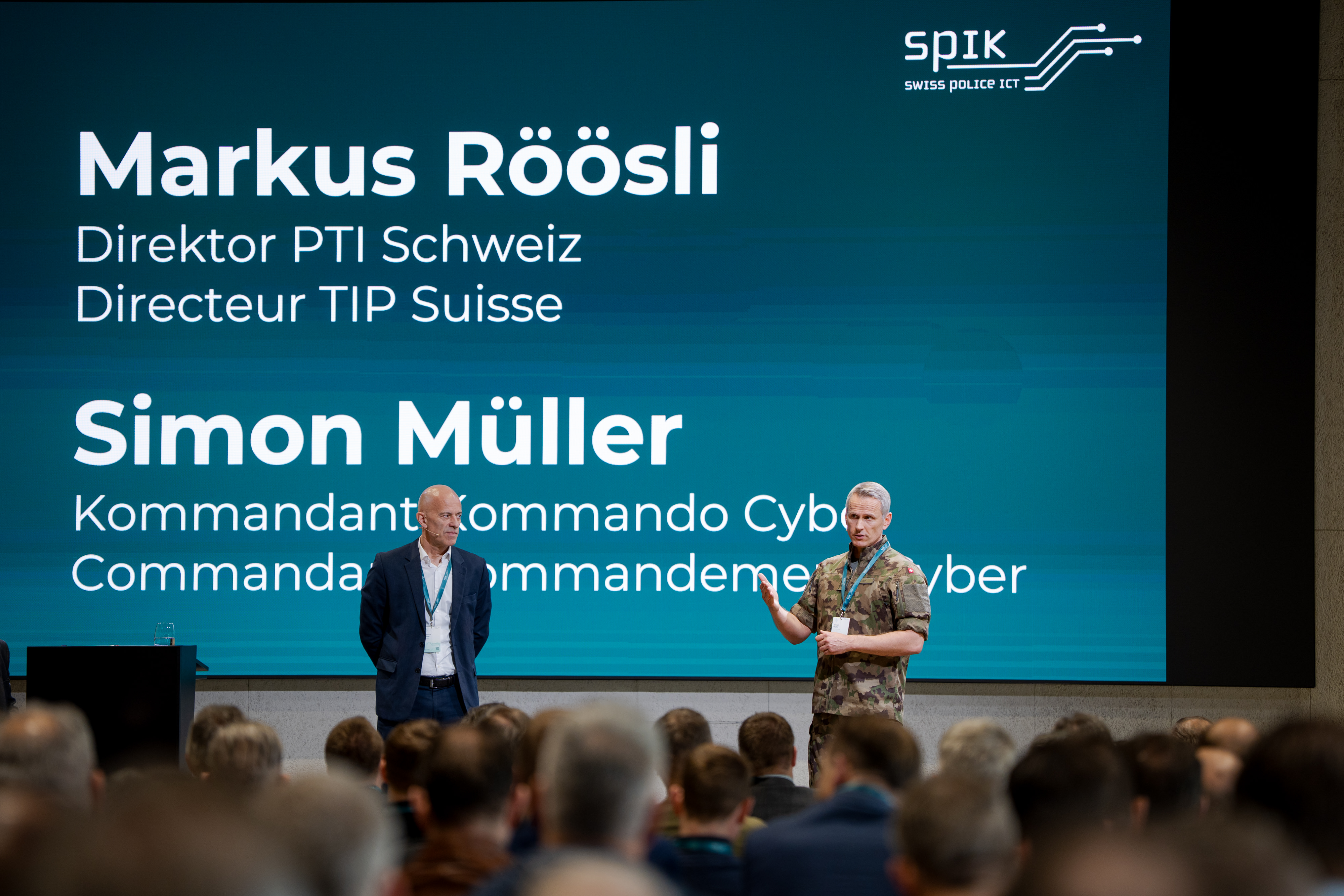 Zwei Männer stehen auf einer Konferenzbühne. Der eine in Militäruniform spricht, der andere im Anzug hört zu. Hinter ihnen eine grosse Leinwand mit Text, davor die unscharfen Köpfe des Publikums.