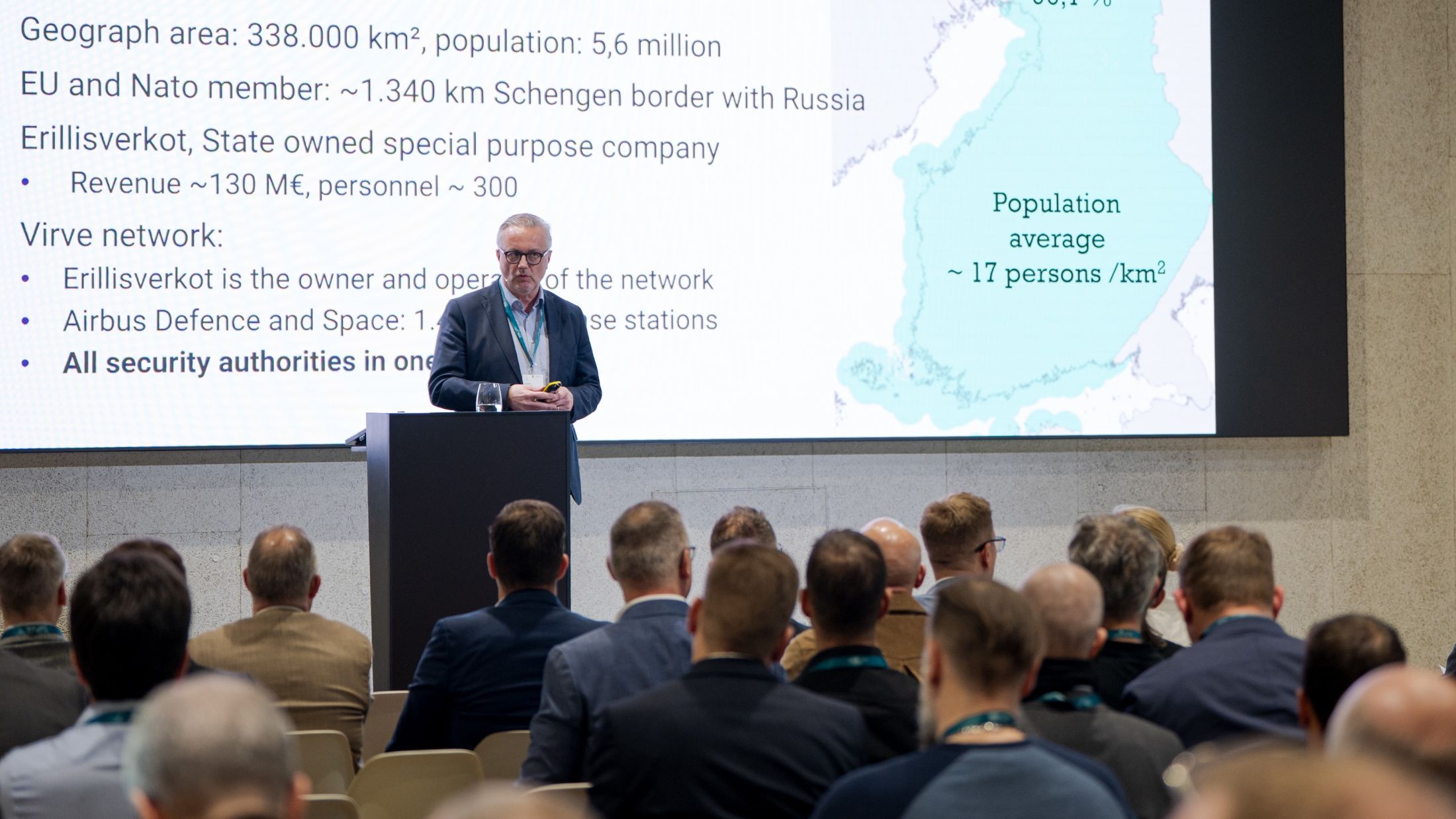 Ein Mann hält auf einer Bühne einen Vortrag. Hinter ihm eine grosse Leinwand mit einer Präsentation über Finnland. Im Vordergrund sind die Hinterköpfe des Publikums zu sehen, das in Reihen sitzt und zuhört.