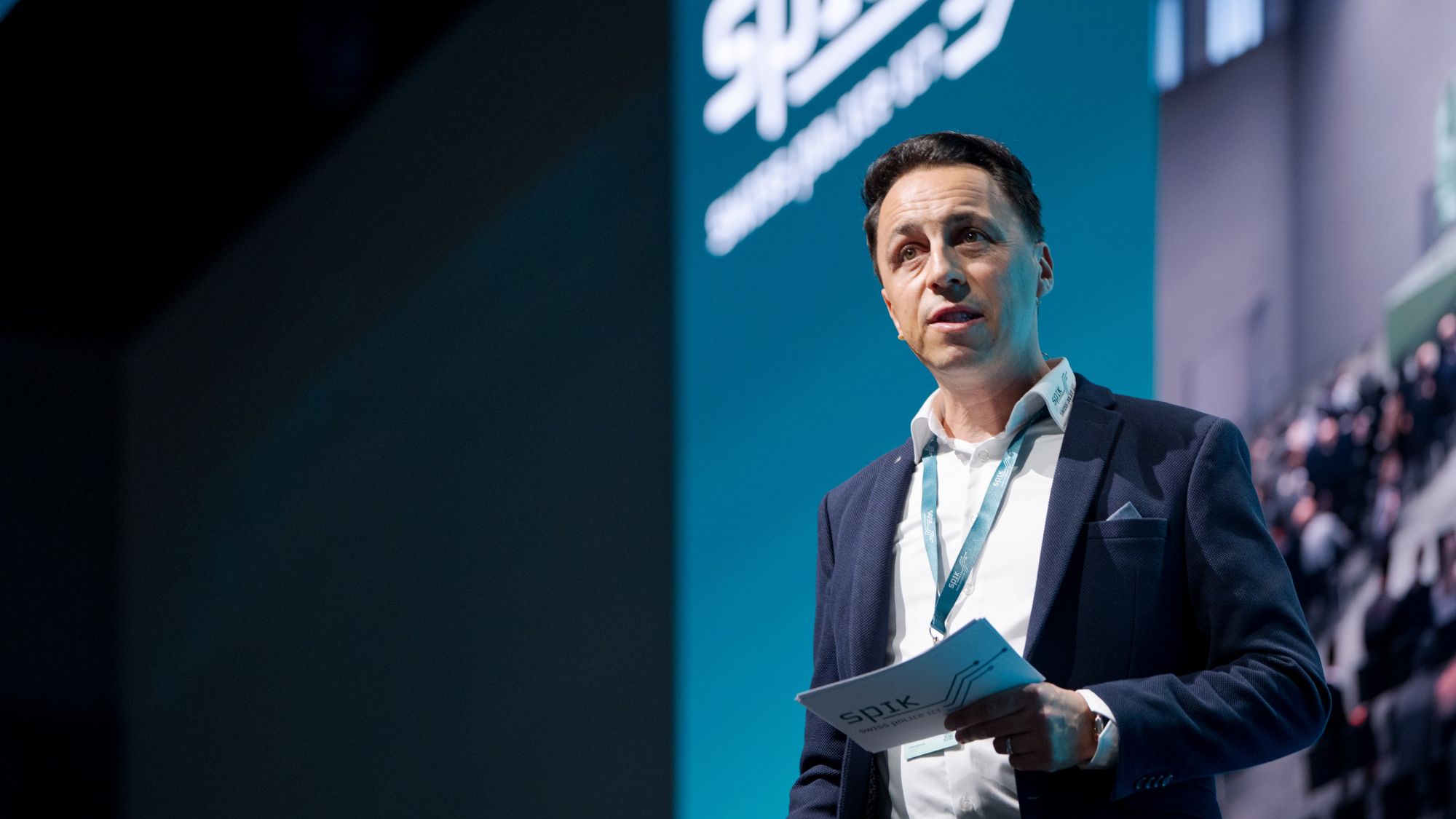 Ein Mann spricht auf einer Konferenzbühne. Er hat dunkles Haar und trägt ein dunkelblaues Sakko über einem weissen Hemd. In der Hand hält er Papiere. Hinter ihm ist eine grosse, türkisfarbene Leinwand und ein Teil des Publikums.