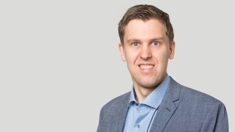 Das Bild zeigt einen jungen Mann mit kurzen Haaren und grauem Anzug vor einem neutral-grauen Hintergrund.