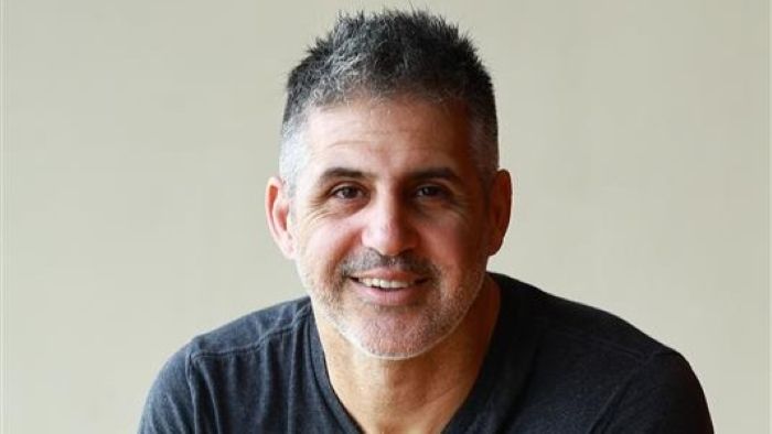 Das Bild zeigt einen lächelnden jungen Mann, der ein dunkles T-Shirt trägt und sich auf ein hölzernes Pult stützt.