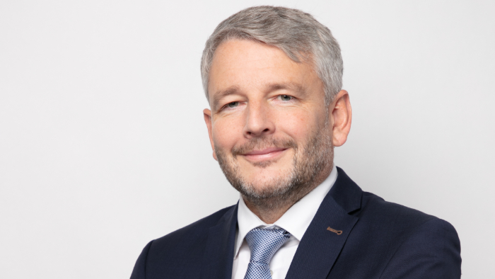 Christopher Tighe blickt freundlich in die Kamera. Es ist ein Porträtfoto mit neutralem Hintergrund.