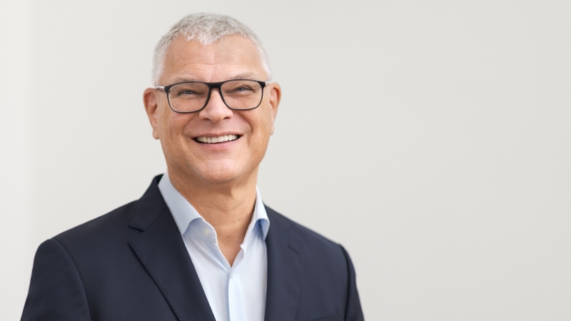 Das Bild zeigt einen lächelnden Mann mit kurzen grauen Haaren und dunkler Brille vor einem hellen Hintergrund. Er trägt ein dunkelblaues Sakko über einem offenen, hellblauen Hemd.